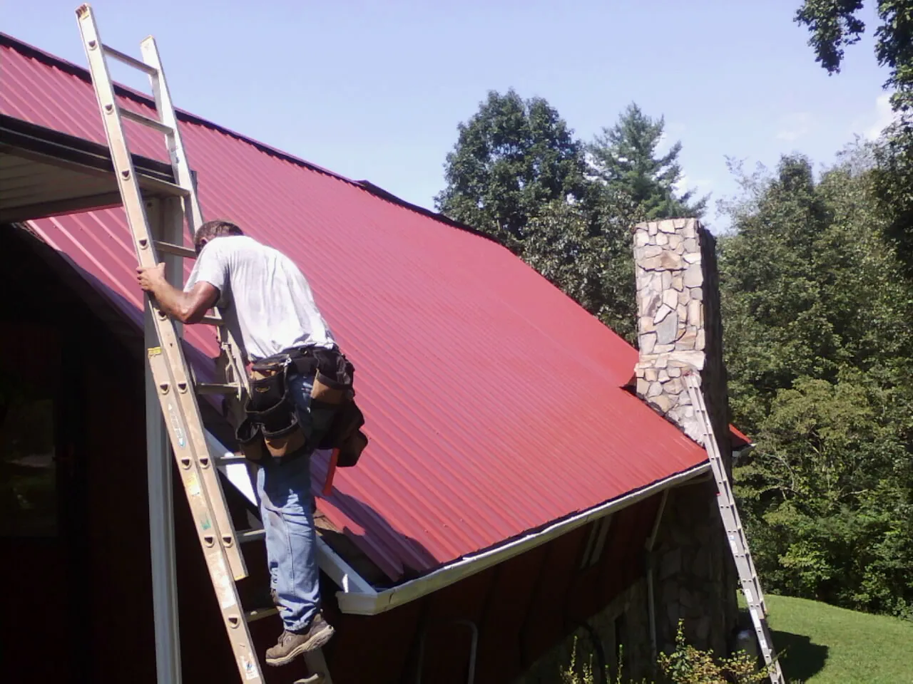 Roofer on a ladder beside a red metal roof and stone chimney — Soffit & Fascia Repair by Roofing Brothers in Cornelius